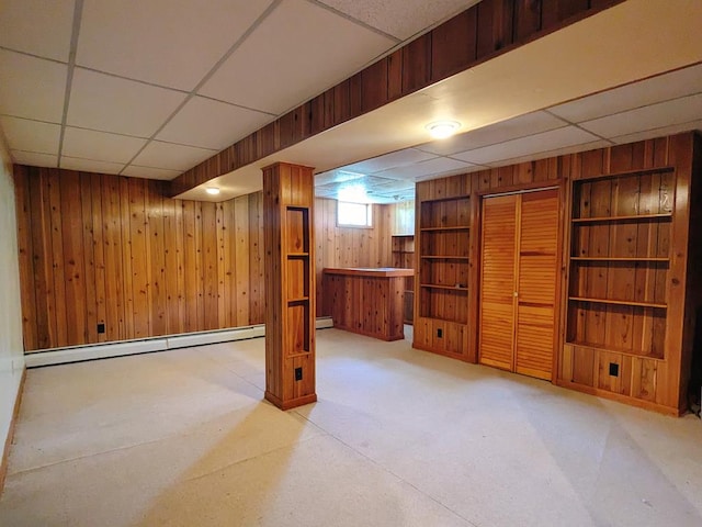 basement with a drop ceiling, wooden walls, and baseboard heating