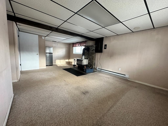 basement with carpet, stainless steel fridge, a drop ceiling, a baseboard radiator, and a wood stove