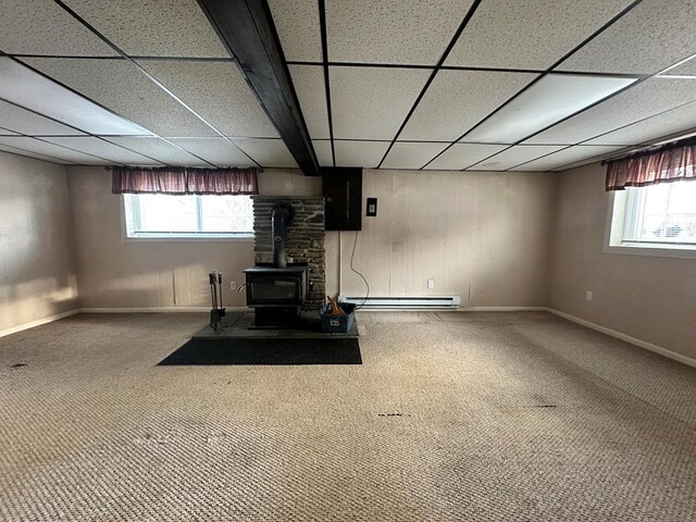 basement featuring carpet floors, a baseboard radiator, a wood stove, and a drop ceiling