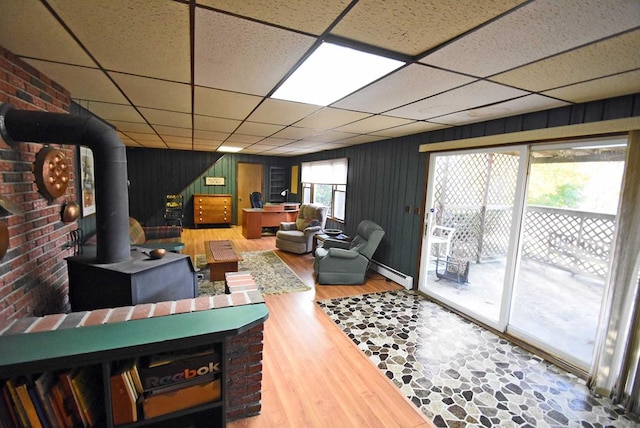 living room featuring a paneled ceiling, baseboard heating, hardwood / wood-style floors, wooden walls, and a wood stove