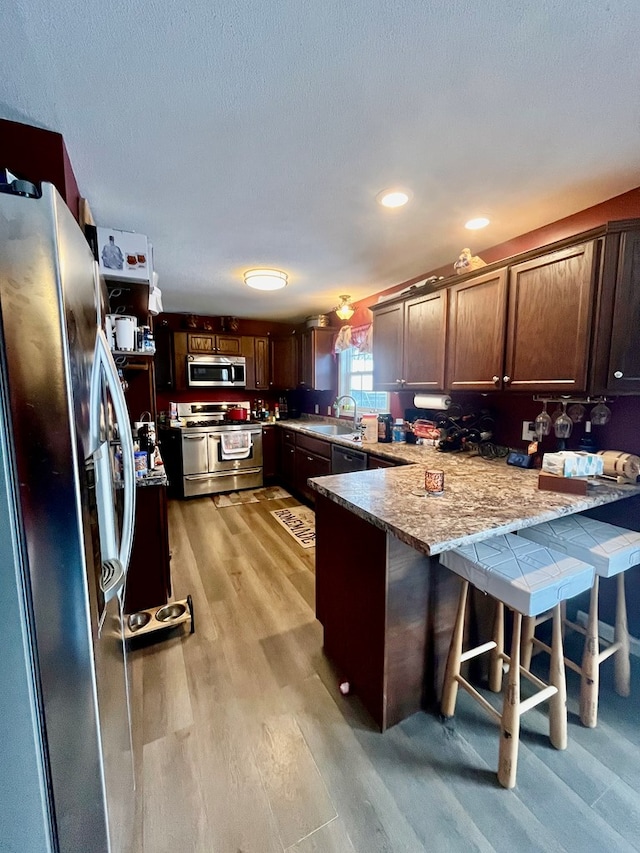 kitchen with kitchen peninsula, appliances with stainless steel finishes, a kitchen bar, light stone countertops, and light hardwood / wood-style floors