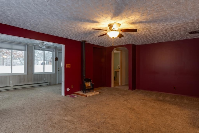 carpeted empty room with a textured ceiling, ceiling fan, a baseboard radiator, and a wood stove