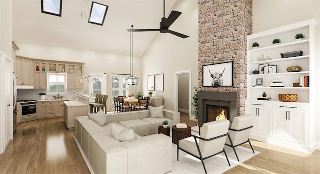 living room with high vaulted ceiling, a stone fireplace, a skylight, ceiling fan, and light wood-type flooring