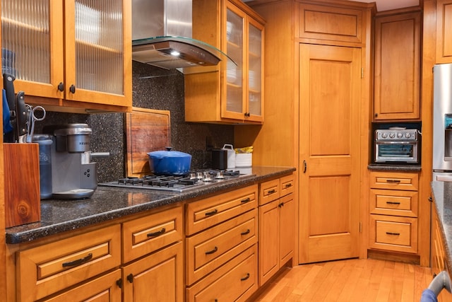 kitchen featuring range hood, dark stone countertops, backsplash, light hardwood / wood-style floors, and stainless steel appliances