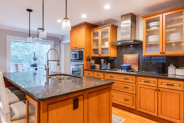 kitchen with wall chimney exhaust hood, sink, dark stone counters, a kitchen island with sink, and backsplash