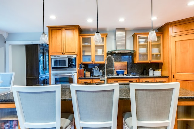 kitchen featuring wall chimney exhaust hood, a kitchen breakfast bar, pendant lighting, stainless steel appliances, and backsplash