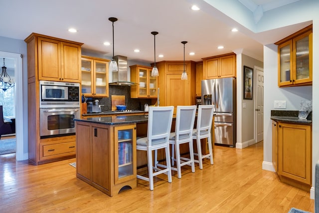 kitchen with a kitchen island, appliances with stainless steel finishes, dark stone countertops, hanging light fixtures, and wall chimney range hood