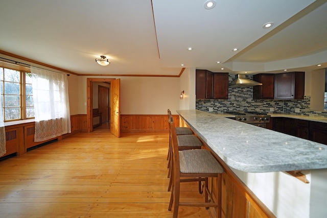 kitchen with wall chimney range hood, a breakfast bar area, high end stainless steel range oven, light hardwood / wood-style floors, and decorative backsplash