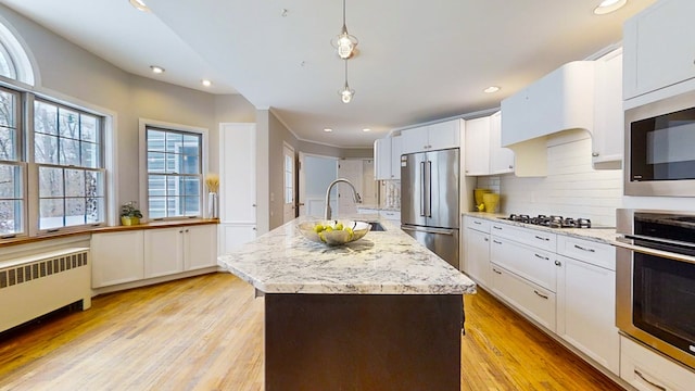 kitchen with appliances with stainless steel finishes, radiator heating unit, pendant lighting, white cabinets, and a kitchen island with sink