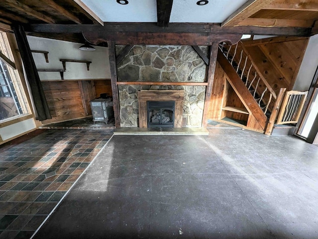 unfurnished living room featuring beam ceiling, wooden walls, a stone fireplace, and a wood stove