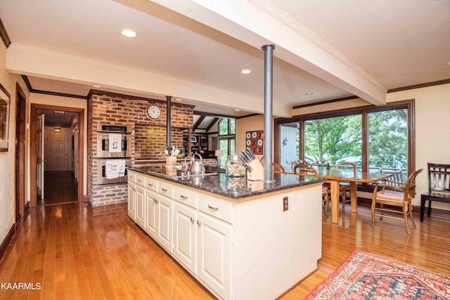 kitchen featuring an island with sink, stainless steel double oven, dark stone countertops, white cabinets, and beamed ceiling