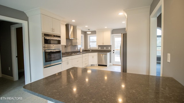 kitchen with stainless steel appliances, wall chimney range hood, white cabinets, dark stone countertops, and sink