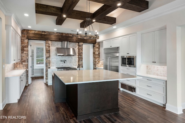 kitchen featuring an island with sink, built in appliances, white cabinets, wall chimney range hood, and tasteful backsplash