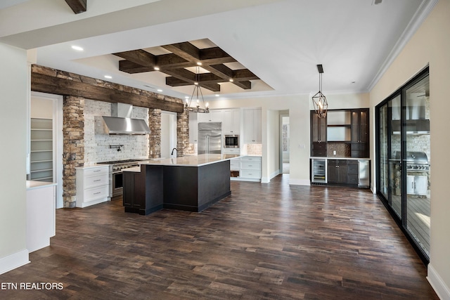 kitchen featuring built in appliances, a center island with sink, coffered ceiling, wall chimney exhaust hood, and white cabinetry