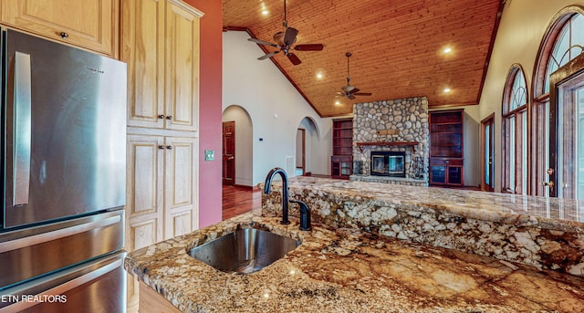 kitchen with ceiling fan, a stone fireplace, stainless steel fridge, high vaulted ceiling, and sink