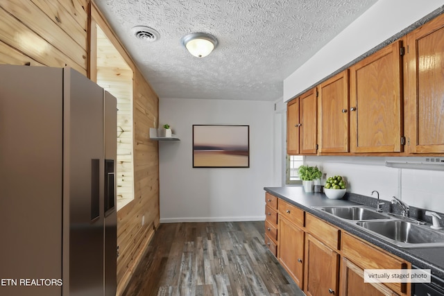 kitchen with a textured ceiling, wooden walls, dark wood-type flooring, stainless steel fridge, and sink