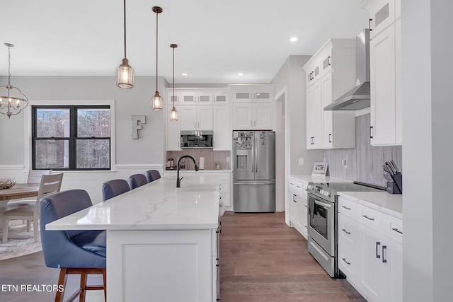 kitchen with a chandelier, a kitchen bar, wall chimney exhaust hood, appliances with stainless steel finishes, and hanging light fixtures