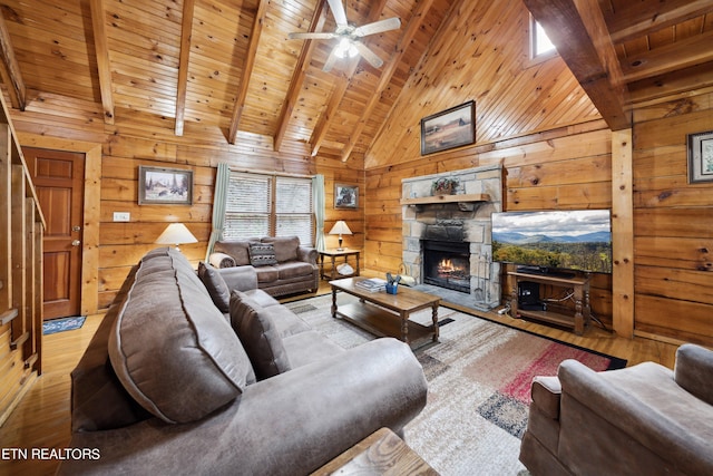 living room with beam ceiling, wood ceiling, a stone fireplace, light hardwood / wood-style floors, and ceiling fan