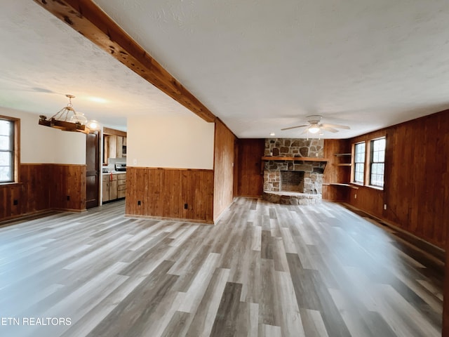 unfurnished living room featuring light wood-type flooring, a textured ceiling, ceiling fan with notable chandelier, beamed ceiling, and a stone fireplace