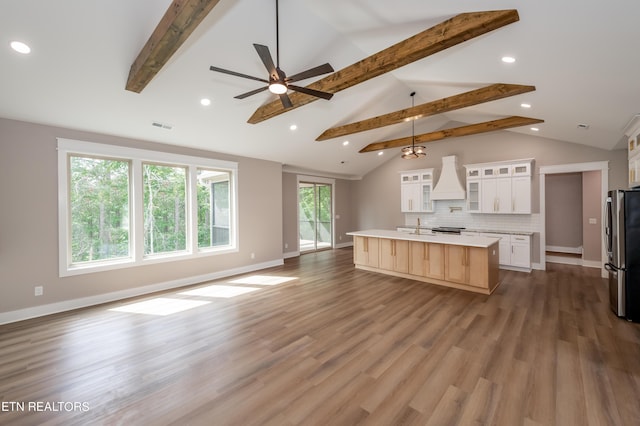 kitchen with a center island with sink, stainless steel fridge, tasteful backsplash, custom range hood, and white cabinetry