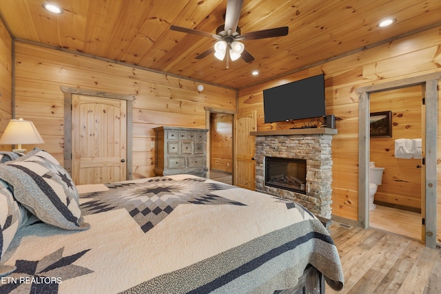 bedroom with light wood-type flooring, wooden ceiling, connected bathroom, a stone fireplace, and ceiling fan