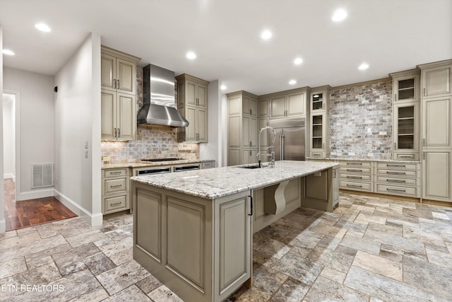 kitchen with stainless steel appliances, decorative backsplash, wall chimney exhaust hood, sink, and a kitchen island with sink