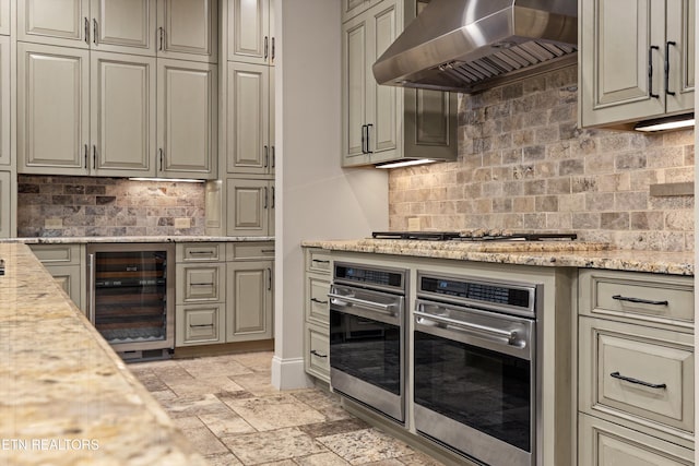 kitchen featuring tasteful backsplash, wine cooler, wall chimney range hood, appliances with stainless steel finishes, and light stone countertops