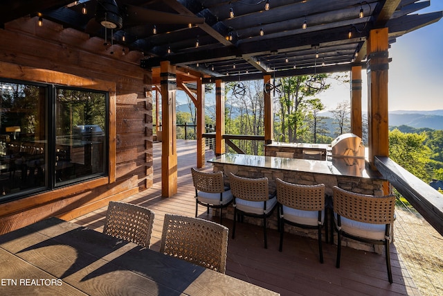 wooden terrace with a grill, an outdoor kitchen, an outdoor bar, a mountain view, and a pergola