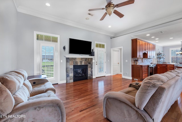 living room with crown molding, ceiling fan with notable chandelier, wood-type flooring, and a stone fireplace