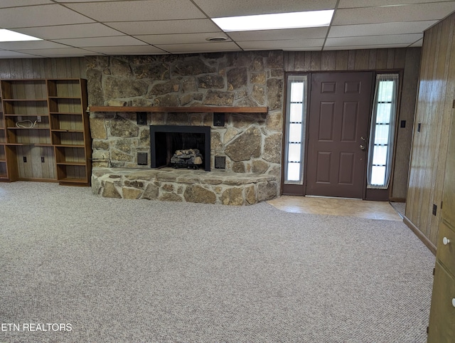 unfurnished living room with carpet floors, a paneled ceiling, wood walls, and a stone fireplace