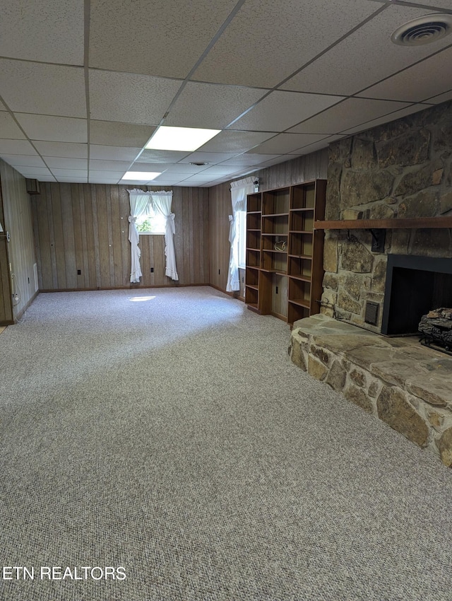 basement with light carpet, wood walls, a drop ceiling, and a stone fireplace