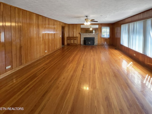 unfurnished living room featuring ceiling fan, a stone fireplace, wood walls, hardwood / wood-style floors, and a textured ceiling