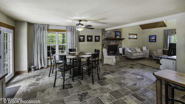 dining area with a textured ceiling, ceiling fan, vaulted ceiling, and a stone fireplace