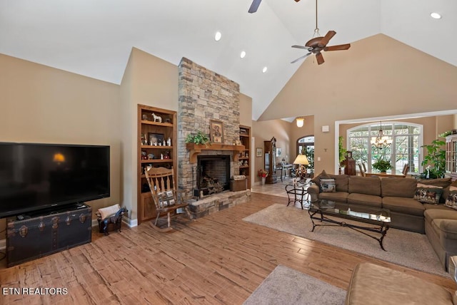 living room with light hardwood / wood-style flooring, built in features, a stone fireplace, high vaulted ceiling, and ceiling fan with notable chandelier