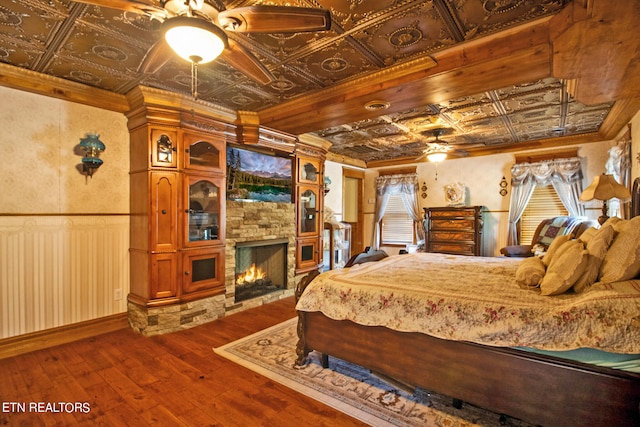 bedroom with crown molding, a stone fireplace, hardwood / wood-style floors, and ceiling fan