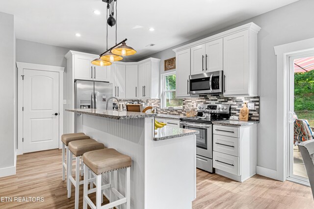 kitchen with appliances with stainless steel finishes, a kitchen island with sink, hanging light fixtures, light stone countertops, and white cabinets