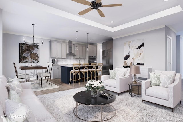 living room with sink, crown molding, light wood-type flooring, a tray ceiling, and ceiling fan with notable chandelier