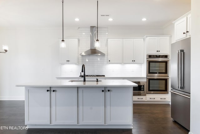 kitchen featuring appliances with stainless steel finishes, white cabinetry, hanging light fixtures, a kitchen island with sink, and wall chimney range hood