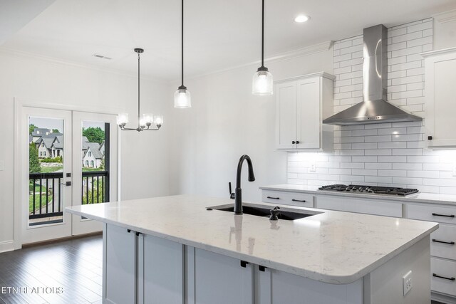 kitchen featuring wall chimney exhaust hood, stainless steel gas cooktop, an island with sink, light stone countertops, and white cabinets