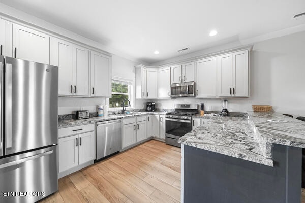 kitchen with appliances with stainless steel finishes, kitchen peninsula, white cabinets, light stone countertops, and light hardwood / wood-style floors