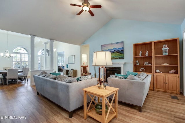 living room featuring high vaulted ceiling, light wood-type flooring, ceiling fan with notable chandelier, and ornate columns