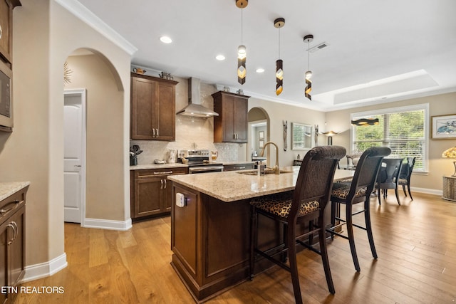 kitchen with arched walkways, wall chimney exhaust hood, a raised ceiling, an island with sink, and stainless steel electric range