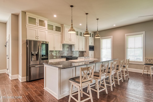 kitchen with a breakfast bar, appliances with stainless steel finishes, glass insert cabinets, an island with sink, and dark stone counters