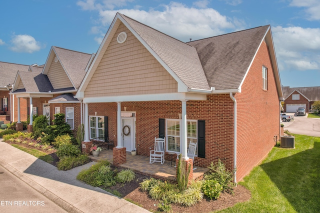 view of front facade featuring central AC unit, a porch, brick siding, roof with shingles, and a front yard