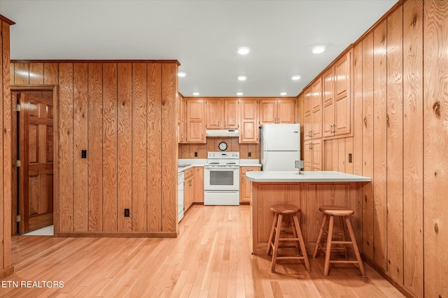 kitchen featuring light hardwood / wood-style flooring, kitchen peninsula, wooden walls, white appliances, and a breakfast bar