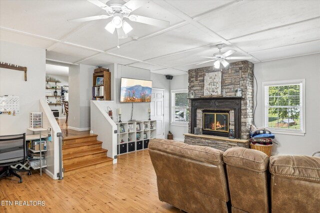 living room featuring ceiling fan, light hardwood / wood-style flooring, a stone fireplace, and coffered ceiling