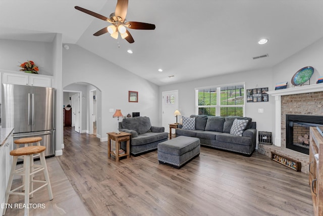 living room with ceiling fan, light wood-type flooring, high vaulted ceiling, and a brick fireplace