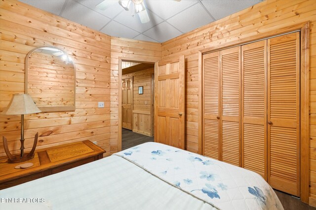 bedroom with a drop ceiling, a closet, dark hardwood / wood-style floors, wooden walls, and ceiling fan