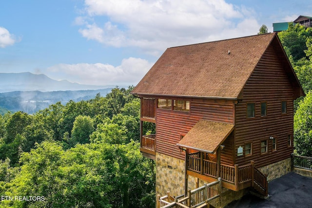 back of house with faux log siding, stone siding, a balcony, a mountain view, and a shingled roof
