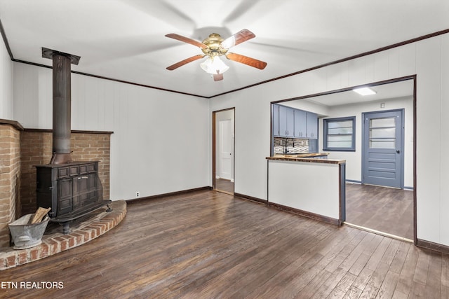 unfurnished living room featuring ornamental molding, a ceiling fan, a wood stove, wood finished floors, and baseboards